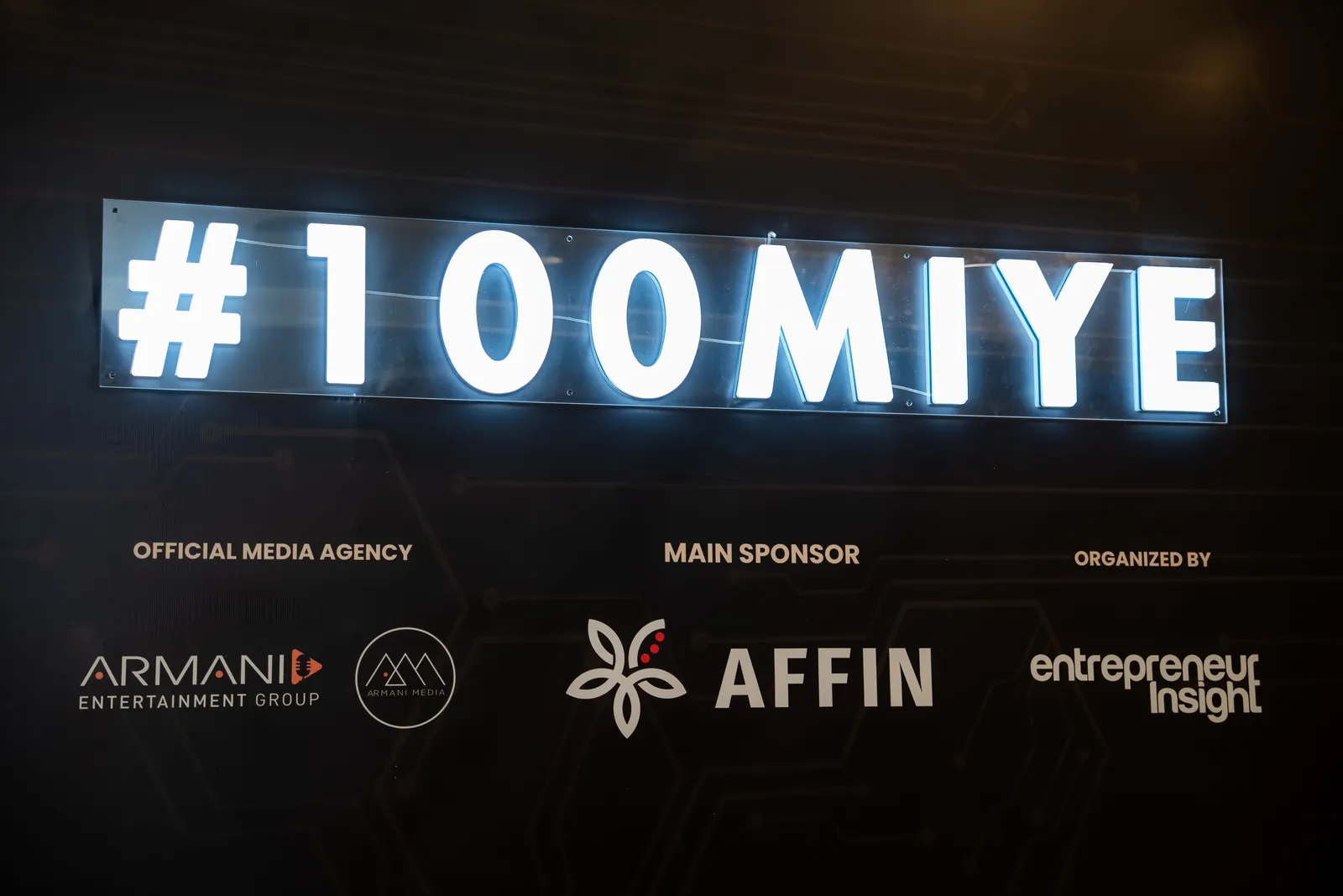 A lit #100MIYE sign above the organiser and sponsor logos