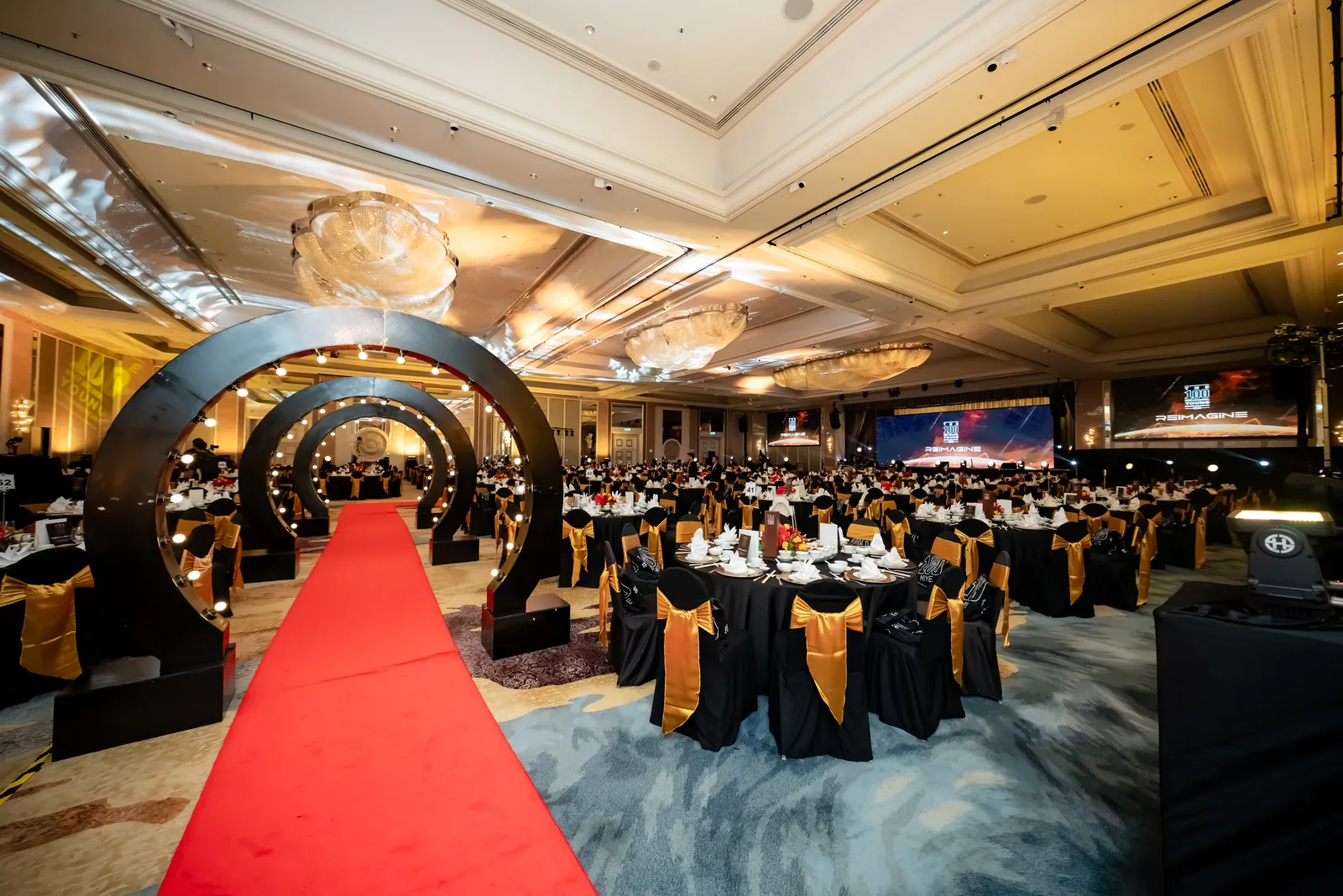 A hotel ballroom set for the 100 MIYE awards, with a red carpet through lit black arches and tables in black and gold
