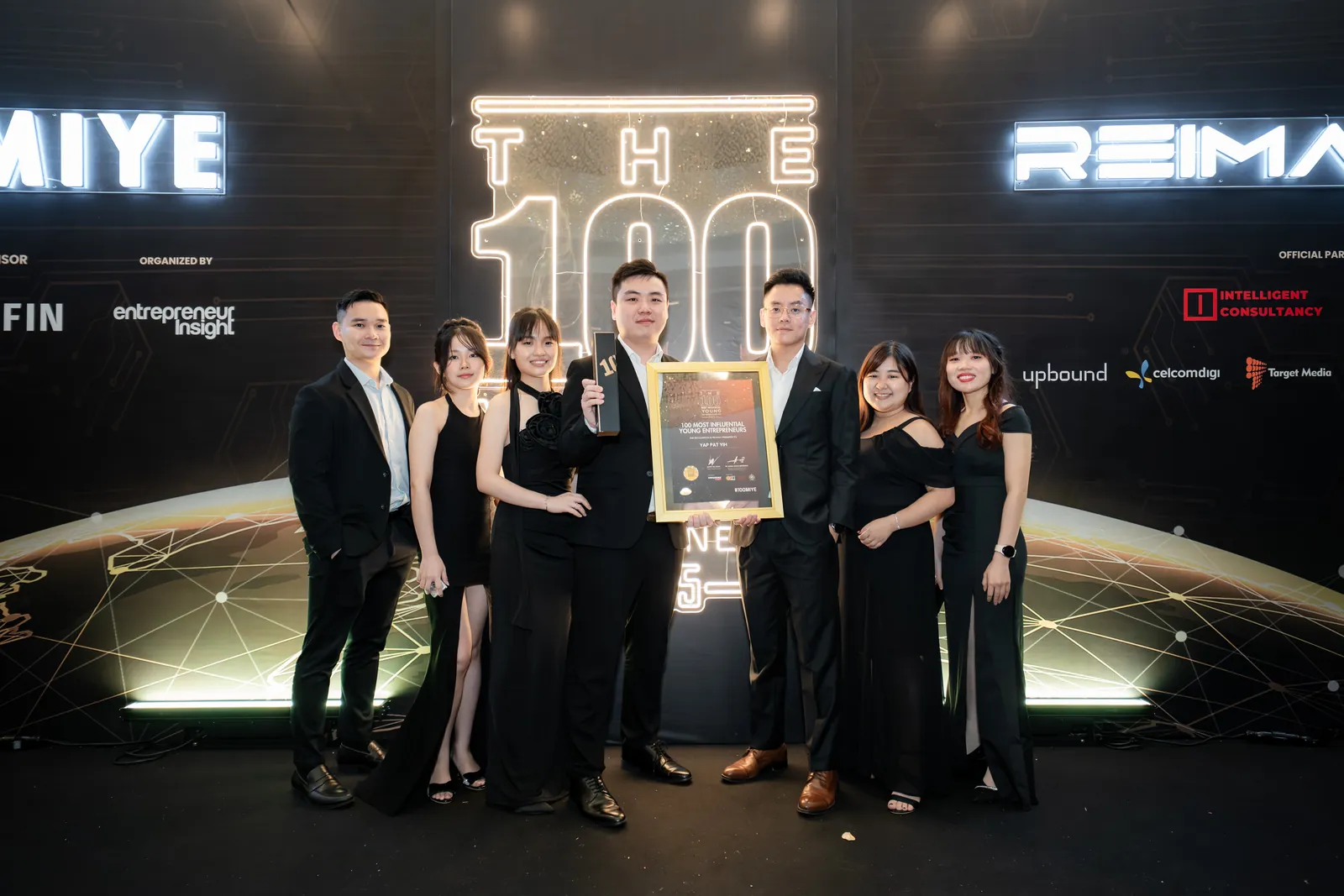 Seven members of the BlackRevo team in front of the 100 MIYE stage backdrop, with Yap Pat Yih in the centre holding the trophy and certificate